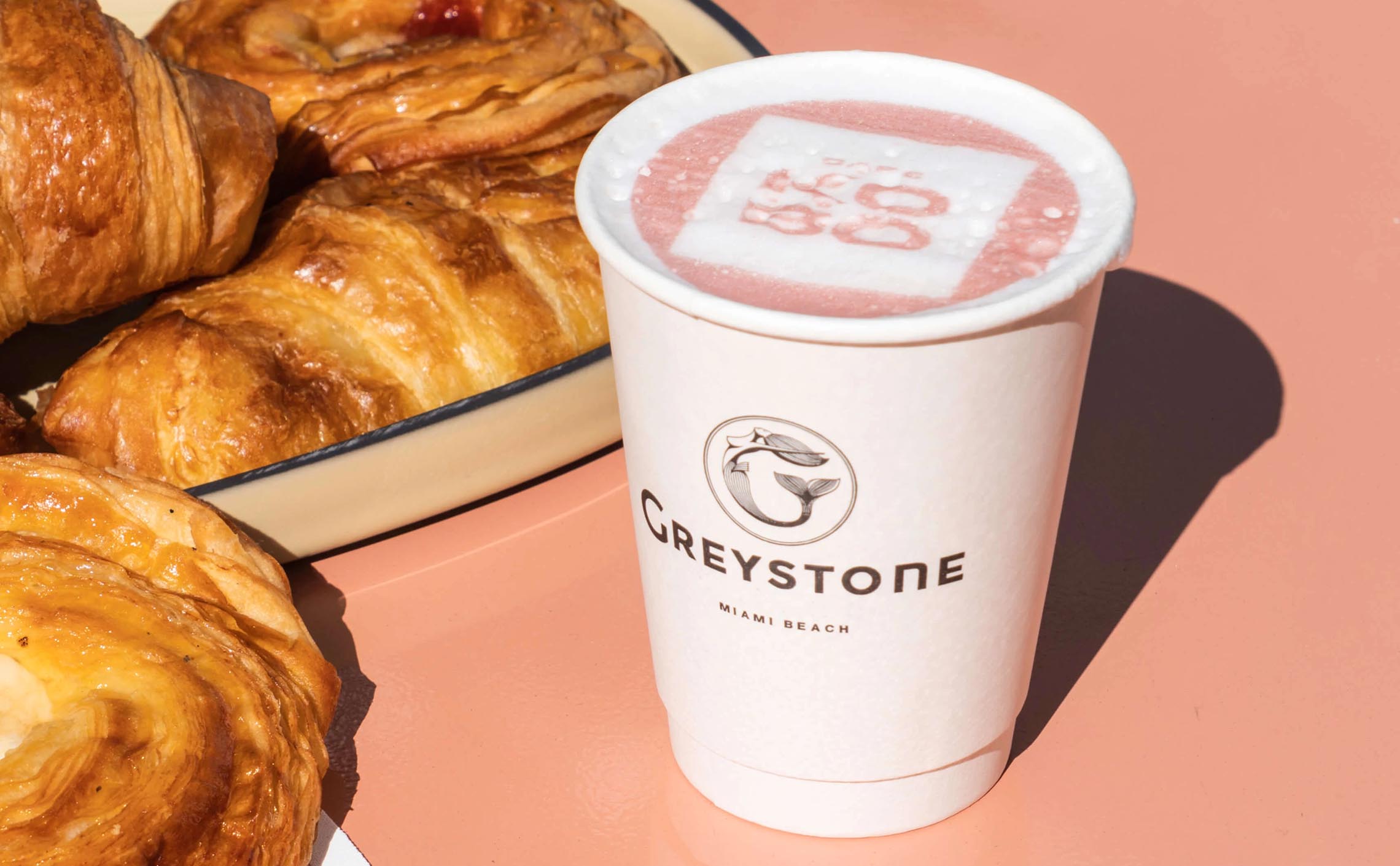Branded paper coffee cup with the Greystone logo with latte foam and a plate of pastries