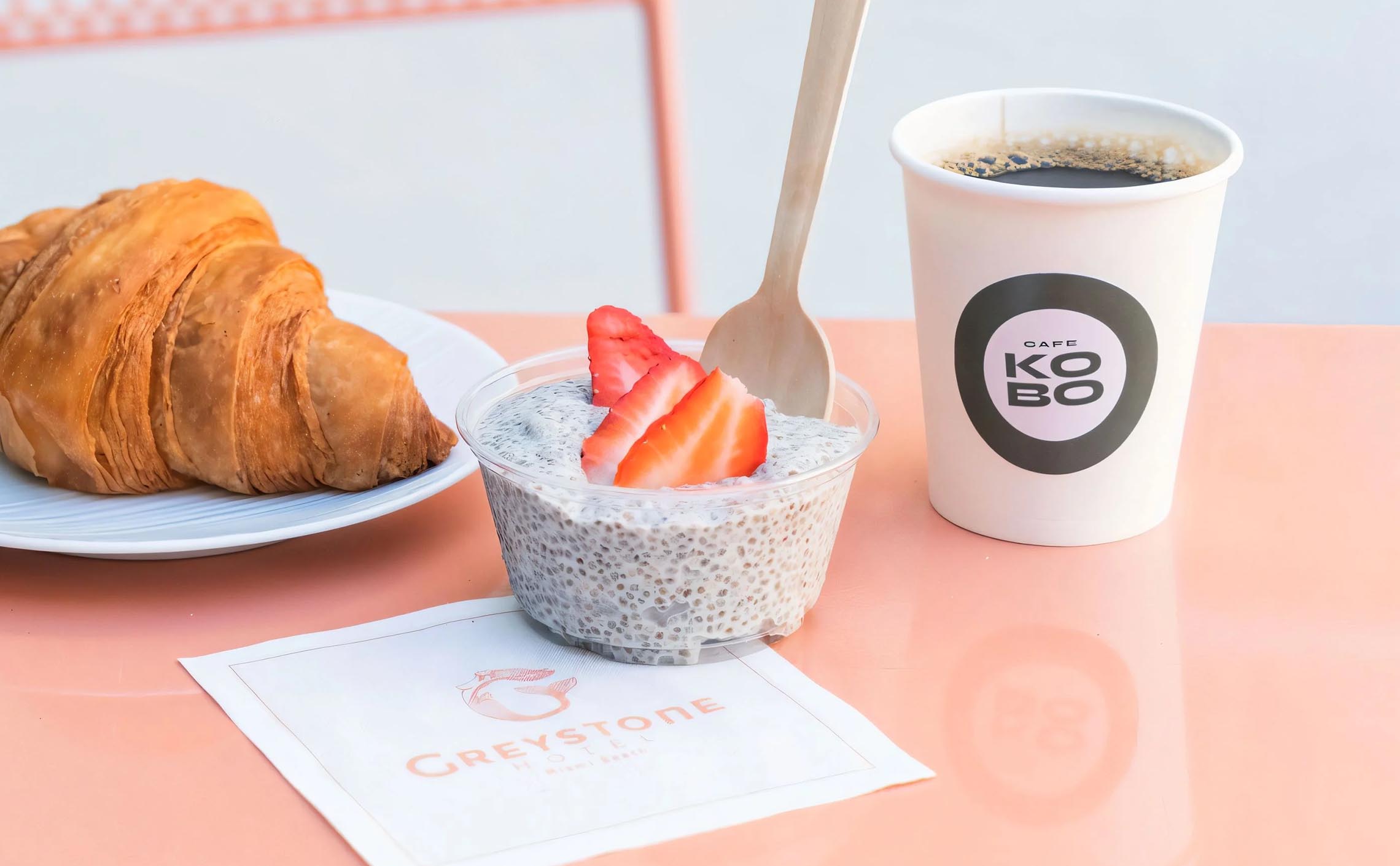 Cjia pudding bowl under a Paper Napkin branded with the Gresytone Logo beside a paper cup of coffee and a fresh croissant